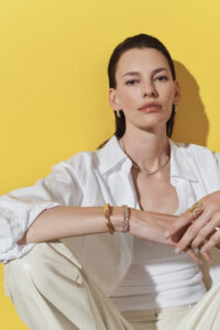 A person with dark hair sits against a bright yellow background, wearing a white shirt and pants. They accessorize with gold jewelry, including earrings, a necklace, bracelets, and a decorative ring. The person’s expression is calm and composed, with relaxed posture and loosely folded arms, showcasing a sense of elegance and understated sophistication.