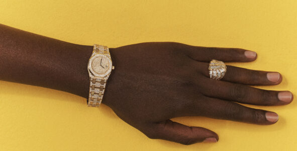 A hand rests on a bright yellow background. It wears an elaborate gold wristwatch with numerous sparkling stones embedded on its surface. The hand is also adorned with a striking gold ring, featuring intricate design work and sparkling embellishments. The overall composition highlights the luxurious jewelry against the bold, contrasting backdrop.