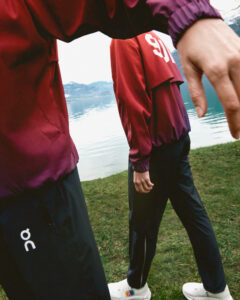 Two individuals wear matching maroon jackets and black pants while walking along a grassy area near a body of water. One arm is extended in the foreground, partially obscuring the view. The landscape features calm water with distant mountains under a cloudy sky, creating a serene outdoor setting that highlights the sporty apparel of the individuals.