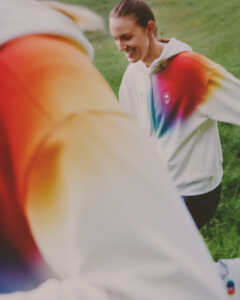 A person with tied-back hair wears a white hoodie that gradually transitions into vibrant shades of red, orange, and blue. They are smiling with eyes directed towards something outside the scene. Another figure, partially visible in the foreground, wears a similar hoodie. The background is a grassy field, suggesting an outdoor setting filled with energy.