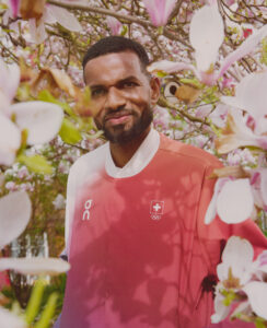 A person with a short beard smiles warmly, surrounded by lush magnolia flowers in full bloom. They wear a red and white shirt, standing amidst the delicate pink and white petals. The scene evokes a sense of springtime renewal and tranquility. Sunlight filters through the branches, casting soft shadows on the serene setting.