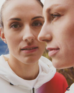 Two individuals stand closely together, both wearing casual hooded jackets. One person faces forward, while the other is in profile, creating a sense of depth and connection. The background hints at an outdoor setting, possibly with natural scenery. The overall mood is calm and contemplative, with subtle expressions suggesting a shared understanding or moment between them.