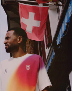 A person with short hair and a beard is wearing a casual shirt with a colorful gradient pattern. Behind, a red flag with a white cross hangs above a building, featuring typical European architectural details. The structure has a mix of textures and colors, including a blue awning. The sky is clear, suggesting a bright day.