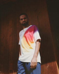 A person stands confidently in front of a wooden background, wearing a bright, gradient-patterned shirt and navy pants. The light catches on their face, highlighting a thoughtful expression. They accessorize with a gold chain and wristband, contributing to a casual yet stylish appearance. The vibrant clothing contrasts with the rustic setting, creating a dynamic visual composition.