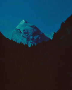 A towering snow-capped mountain dominates the scene, bathed in a deep blue twilight. The impressive peak stands prominently against the dark silhouettes of surrounding trees. The serene, atmospheric landscape conveys a sense of majesty and tranquility, with the icy summit starkly contrasting the shadowed forest below. The sky transitions smoothly from rich blue to the darkened horizon.