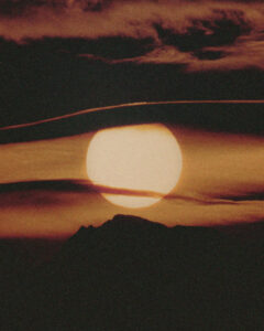 A large, glowing sun sets on the horizon, casting a warm, golden hue across the sky. The silhouette of a mountain is visible below, while dark, wispy clouds drift across the scene. The rich, deep colors create a dramatic contrast, highlighting the serene beauty and tranquility of nature as daylight fades into dusk.