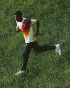 A person with short hair is running energetically across a grassy field. They are wearing a white shirt with a vibrant gradient pattern, along with dark pants and white shoes. The natural surroundings are lush and green, emphasizing a sense of motion and vitality. The scene conveys a dynamic and active outdoor experience.