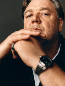 A contemplative man gazes intently, resting his chin on folded hands. His expression is thoughtful, with furrowed brows suggesting deep reflection. He wears a dark jacket over a white shirt, and his left wrist sports a metallic watch. A ring adorns one of his fingers. The background is softly blurred, emphasizing his introspective demeanor.
