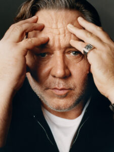 A middle-aged person with light facial stubble and expressive eyes holds their head with both hands, fingers interlaced across their forehead. The intense gaze and slightly furrowed brow suggest deep thought or contemplation. They wear a dark jacket over a white shirt, and a prominent silver ring adorns one finger, adding a touch of subtle elegance.