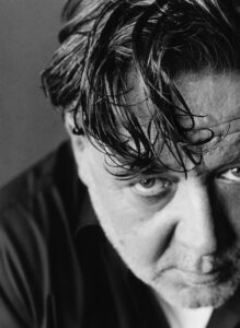 A close-up in black and white reveals a person with wet hair falling over their forehead, partially obscuring intense, thoughtful eyes. The face shows signs of age with a slight stubble and wrinkles. The top of a dark shirt is visible, suggesting a somber or contemplative mood, enhanced by the sharp contrast and lighting.