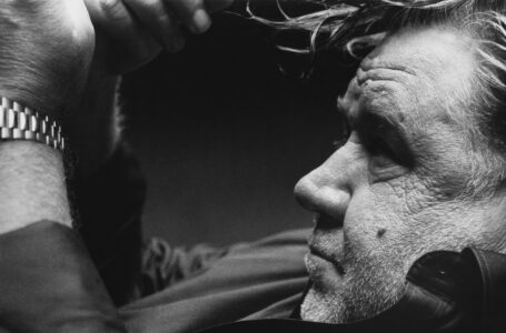 A grayscale close-up captures an older man with a focused expression. His wrinkled forehead and stubbly beard highlight his mature features. Loose strands of hair fall over his face as he rests against a violin's chin rest. A metallic watch wraps his wrist. Shadows accentuate the contemplative mood, while the background remains soft and unobtrusive.