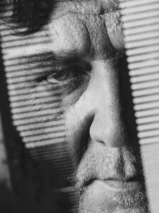 A close-up reveals a man's face partially obscured by blinds, casting striped shadows. His eyes appear intense, emphasizing a sense of contemplation or mystery. The texture of his skin and subtle details, like facial hair, add depth to the scene. The monochromatic tones reinforce a somber, introspective mood, evoking curiosity about the man's thoughts.
