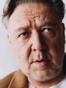 A middle-aged man with short, light brown hair and a slightly rugged face gazes intently. His facial hair is trimmed, and he wears an open-collared brown coat over a white shirt. The close-up view highlights his textured skin and expressive eyes, giving a sense of introspection. The background is softly blurred, emphasizing his features.