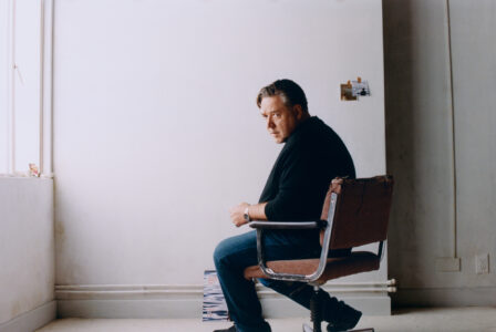 A man sits pensively on a swivel chair in a minimalist room with a neutral color scheme. Soft light filters through a window on the left, illuminating the scene. The walls are bare, except for two small, colorful pictures affixed near the corner. Sparse furnishings and an uncluttered floor contribute to the tranquil, contemplative atmosphere.
