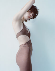 A person with short, curly hair poses against a pale background, wearing a form-fitting brown outfit with cut-out sections. One arm is raised, revealing a sleek silhouette and a contemplative expression. The minimalist setting and soft lighting highlight the texture and color of the clothing, creating a serene and stylish ambiance.