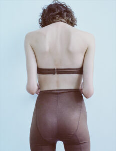 A person with curly hair is viewed from behind, standing against a plain light background. They are wearing a strapless top and high-waisted pants, both in a similar shade of brown. The posture shows relaxed shoulders, and the overall atmosphere has an understated, minimalistic feel, emphasizing simplicity and naturalness.