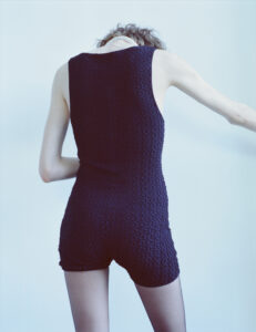 A person with short, tousled hair is wearing a textured black sleeveless outfit. The figure is captured from the back, with their arms slightly extended. The lighting creates a cool ambiance, accentuating the texture of the garment. The overall setting evokes a minimalist and modern aesthetic, with a focus on simplicity and style.