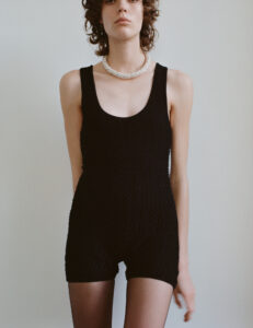 A person stands poised wearing a textured black sleeveless jumpsuit. Their hair is softly curled, and they adorn a statement piece, a large, elegant pearl necklace. The minimalistic background enhances the simplicity and sophistication of the outfit, drawing focus to the person's confident posture and the intricate detailing of their ensemble.