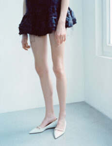 A person stands on a light gray floor, with white pointed shoes. They're wearing a dark, layered, frayed mini dress that contrasts with the minimalist surroundings. Their lightly tanned skin is visible from the thighs down, and their arms hang naturally by their sides. Natural light enters from the right, suggesting a window nearby, adding a soft glow.