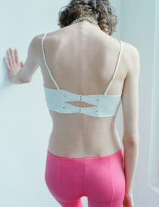 A person stands with their back facing the observer, gently leaning against a white surface. They wear a white strap top with intricate fastening and bright pink pants. The person’s shoulder-length hair curls slightly, and they have delicate jewelry around their neck. The soft natural light enhances the minimalist and serene atmosphere, highlighting the details and textures of the clothing.