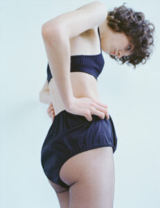 A person with curly hair stands in a soft, minimal setting, wearing dark undergarments. They are bent slightly forward, with hands on their hips, exhibiting a thoughtful pose. The background is a muted, pale color, enhancing the focus on their posture and attire. The lighting is natural, gently highlighting the texture of the clothing and skin.