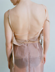 A person stands with their back to the viewer, wearing a delicate, sheer dress. The garment features thin straps and flows elegantly downwards. The skin is illuminated softly, showcasing a gentle natural tone. The overall scene exudes serenity and subtlety, highlighting the intricate texture and design of the fabric against the person’s bare skin.