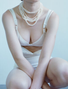 A person is seated with arms crossed, wearing a white outfit and several layers of pearl necklaces. The focus is on the upper body, highlighting the intricate jewelry against simple attire. The background is a soft, light color, enhancing the elegance and minimalism of the composition. The posture suggests a relaxed yet poised demeanor.