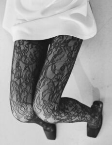 A person is seated, one leg crossed over the other, displaying intricate lace-patterned hosiery in black, revealing floral and swirling designs. The upper part of their attire is a simple, smooth fabric of a light color, contrasting with the texture below. They are wearing pointed, dark shoes, enhancing the overall elegant and fashionable appearance. The angle emphasizes the textiles and patterns.