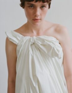 A person with short brown hair gazes directly ahead, exuding a calm and introspective demeanor. They have light skin and their attire is a loosely draped white fabric, gathered at the shoulder into a knot. The background is an unfocused light gray, enhancing the serene and minimalist composition of the scene.