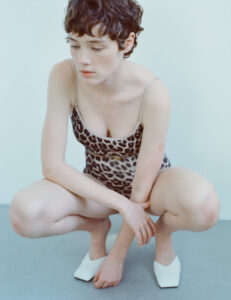 A person with short, dark hair is crouching with a contemplative expression. They are wearing a patterned, sleeveless top and white shoes with pointed toes. The background is a soft, pale blue-gray, creating a calm and minimalist atmosphere. The focus on the individual's relaxed posture and direct gaze evokes a sense of introspection and tranquility.