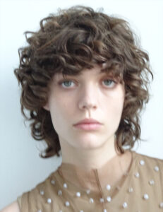 A person with short, curly brown hair is seen on a light background. They have light blue eyes and are wearing a sleeveless garment adorned with small, shiny embellishments. The expression is neutral, with a slightly tilted head. The lighting is soft, highlighting the texture of the hair and the details of the fabric.