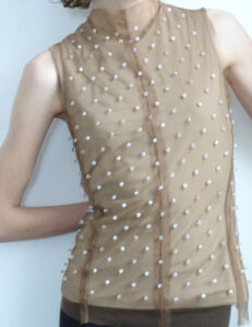 A person is wearing a sleeveless, sheer, tan top adorned with numerous small, round embellishments scattered evenly across the fabric. The top has a high neckline and a central seam running vertically down the front. The person's left arm is bent, with a hand resting on their back, creating a relaxed pose against a plain background.