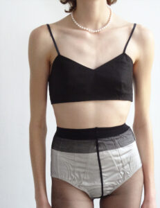A person is wearing a black cropped top with thin straps and sheer high-waisted briefs. A pearl necklace adorns their neck, contrasting with the simplicity of the attire. The background is plain and neutral, enhancing the focus on the clothing and accessories. The style is minimalist and modern, highlighting elegant and vintage-inspired fashion elements.