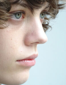 A close-up profile view showcases a youthful face with delicate features: soft, slightly parted lips, a smooth, fair complexion, and vibrant blue eyes gazing intently ahead. The hair cascades in loose curls, adding a touch of playfulness to the serene expression. The background is blurred, emphasizing the contemplative mood and the subject’s introspective focus.