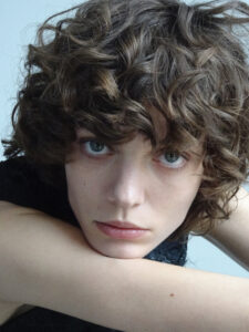 A person with curly, tousled hair gazes directly with a neutral expression. Their head rests on their arm, creating a relaxed pose. The lighting is soft, highlighting their features and the texture of the hair. The background is simple and unobtrusive, allowing focus on the person's thoughtful demeanor and expressive eyes.