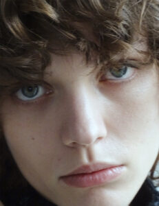 A close-up view showcases a face with curly brown hair framing a fair complexion. The individual has striking blue eyes that gaze directly forward. Soft, natural lighting highlights the smooth texture of the skin and the relaxed, slightly parted lips. The overall expression carries a calm, introspective, and serene quality.