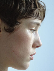 A close-up profile of a person with short, wavy brown hair gazing downward. The skin has a natural texture, highlighted by soft lighting that emphasizes the serene expression. The gentle curve of the nose and the subtle definition of the lips and chin contribute to a contemplative and introspective mood against a smooth, light background.