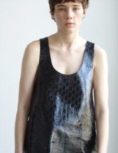 A person with short hair looks directly at the viewer, wearing a shiny sleeveless black top. The material has a textured, embossed pattern. Their expression is neutral, set against a light, unadorned background that enhances the focus on their upper body. The overall mood appears calm and introspective, with a minimalist aesthetic.