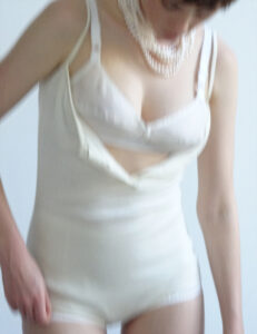 A person is depicted in soft focus, wearing a form-fitting, light-colored undergarment. Their shoulders are draped with a layered pearl necklace, and the outfit includes a strap detail. The pose suggests a downward glance, emphasizing elegance and a minimalist aesthetic. The composition uses faint colors and subtle lighting, creating a dreamy, ethereal effect that evokes timeless sophistication.