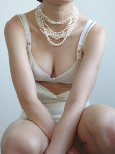 A person is seated with arms crossed over their knees, wearing an off-white bra and matching skirt. Several strands of pearl necklaces gracefully adorn the neck, cascading elegantly. The lighting is soft, emphasizing the smooth skin. The posture is relaxed, and the background is a simple, neutral tone, drawing attention to the jewelry and attire.