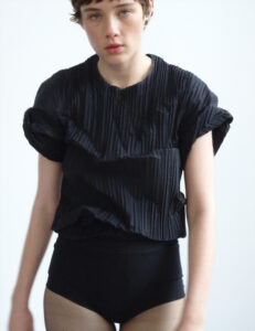 A person gazes forward wearing a black, short-sleeved, textured top paired with matching high-waisted bottoms. The outfit features subtle, vertical pleats, giving it a structured look. The individual has short hair and a neutral expression. The background is simple and plain, emphasizing the focus on the person's appearance and attire, exuding a minimalist and modern style.