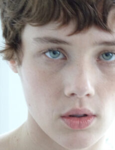 A close-up reveals a person with short, slightly tousled brown hair. Their light blue eyes gaze directly forward, framed by faintly arched eyebrows. The face appears soft, with a calm expression. The lips are slightly parted, showing a hint of natural color. The overall tone feels serene, with soft, diffused lighting enhancing the features.