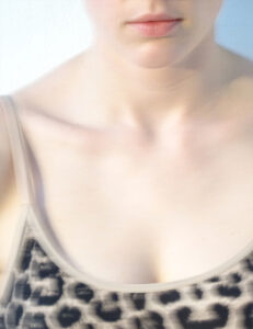 A close-up view captures the lower face and upper body of a person wearing a leopard print top. Soft, natural lighting highlights features such as lightly textured lips and a smooth complexion. The neckline of the garment is scooped, emphasizing the collarbone, and the overall appearance exudes a relaxed, casual vibe. The background is plain and unobtrusive.