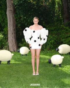 A woman stands on a grassy field surrounded by sheep-shaped sculptures. She wears a short white dress with oversized puffy sleeves and large black polka dots. The backdrop includes lush greenery and trees, creating a serene, whimsical setting. Her hair is tied back, and she is positioned centrally, giving emphasis to her striking outfit and the artful scene.