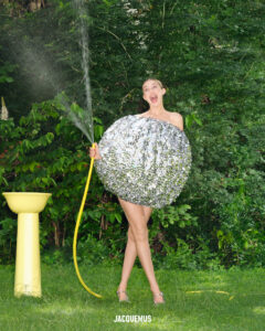 A person stands in a lush garden, joyfully holding a watering hose that sprays water. They wear a large, spherical, shimmering silver outfit, resembling a disco ball. The bright, summer setting includes a yellow, pedestal-like structure nearby, surrounded by vibrant green foliage, creating a playful and whimsical scene.