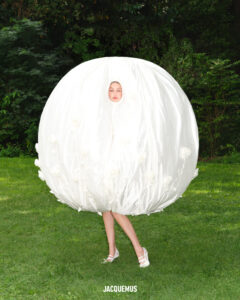 A person is wearing a voluminous, spherical white outfit that covers most of their body, with only their face and legs visible. The setting is outdoors on a lush green lawn with trees and foliage in the background. The ensemble is playful and avant-garde, reflecting a creative, artistic fashion concept that contrasts with the natural environment.