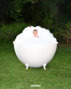 A large, white, round bathtub stands on a lush green lawn, surrounded by trees. A person with a relaxed expression is submerged in bubbles, with only their head visible above the frothy surface. The scene is serene and whimsical, creating a striking contrast between the natural setting and the unusual placement of the bathtub outdoors.
