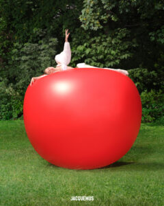 A person in white clothing gracefully poses atop a large, glossy red sphere resembling a cherry, set against a lush backdrop of green grass and dark foliage. The composition contrasts the vivid red with the natural surroundings, creating an artistic and surreal scene that captures a blend of elegance and whimsy.