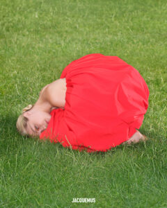 A person is curled up on a lush, green grass lawn, wearing a voluminous, bright red fabric that envelops them almost entirely. Their head is resting sideways, nestled against the grass, with eyes softly focused forward. The vivid contrast between the red fabric and green grass creates a striking and artistic visual scene.