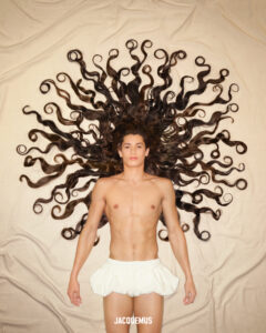 A person lies on their back, eyes closed, on a beige surface. Their long, curly hair fans out dramatically, resembling a series of spirals or waves radiating outward. The individual is shirtless, wearing a short, white, tutu-like skirt. The composition evokes a sense of artistic expression and creativity, with a focus on the intricate design of the hair.